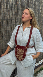 A woman wearing a white linen shirt and pants models a brown leather crossbody pouch with gold embroidery and red straps, set against a bamboo screen background.