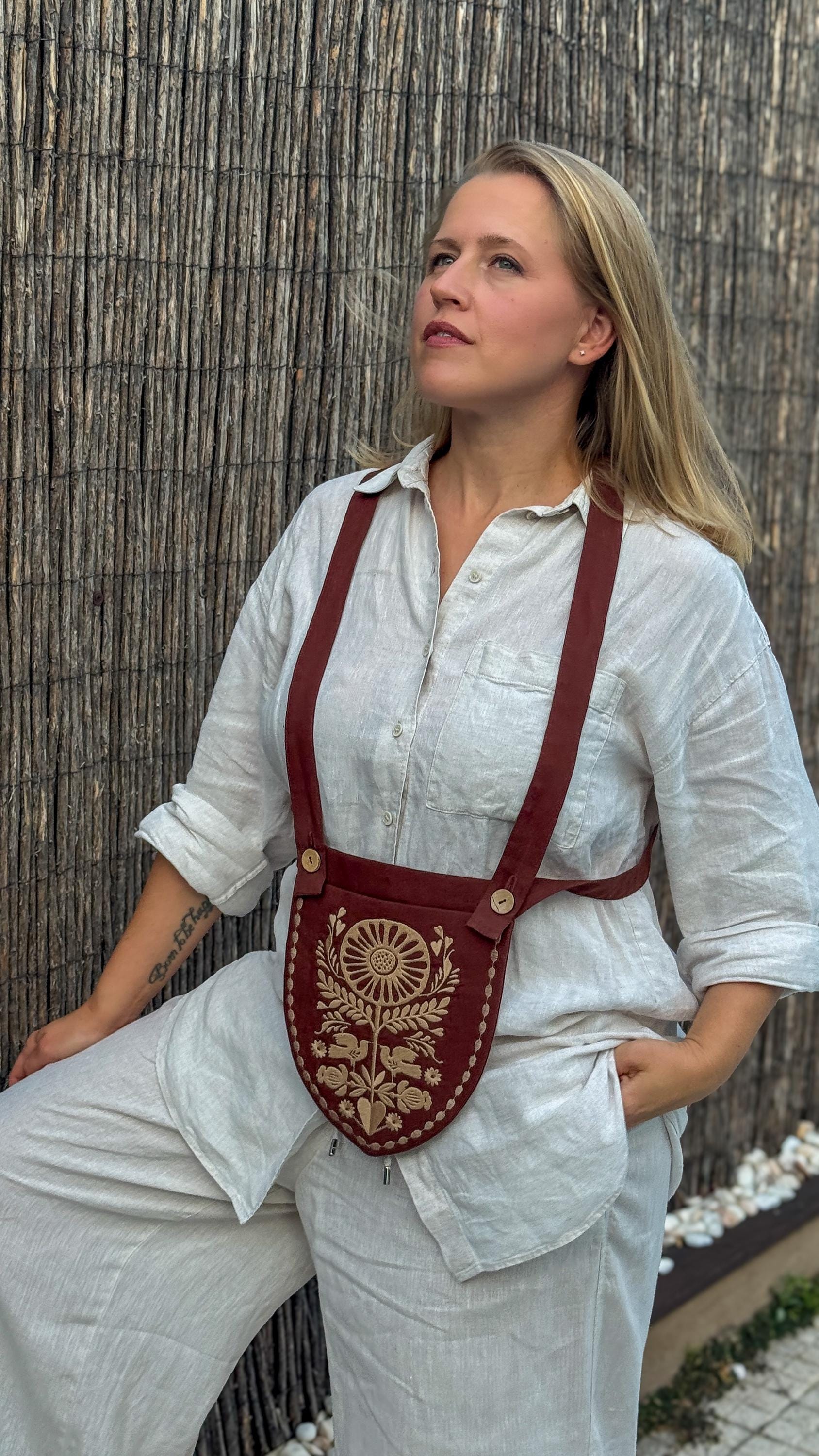A woman wearing a white linen shirt and pants models a brown leather crossbody pouch with gold embroidery and red straps, set against a bamboo screen background.