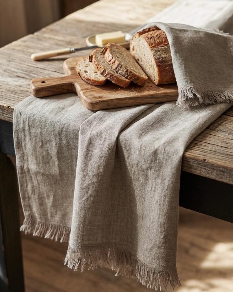 A light beige linen tablecloth with fringed edges draped over a rustic wooden table, styled with a wooden cutting board, sliced artisanal bread, and a butter knife.