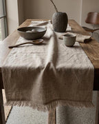 A light beige linen table runner with fringed edges draped over a rustic wooden dining table, styled with ceramic bowls, a wooden spoon, and a textured vase.