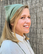 A smiling blonde woman wearing a sage green fabric head covering or bandana, paired with a light-colored collared shirt, standing against a textured bamboo or reed screen.