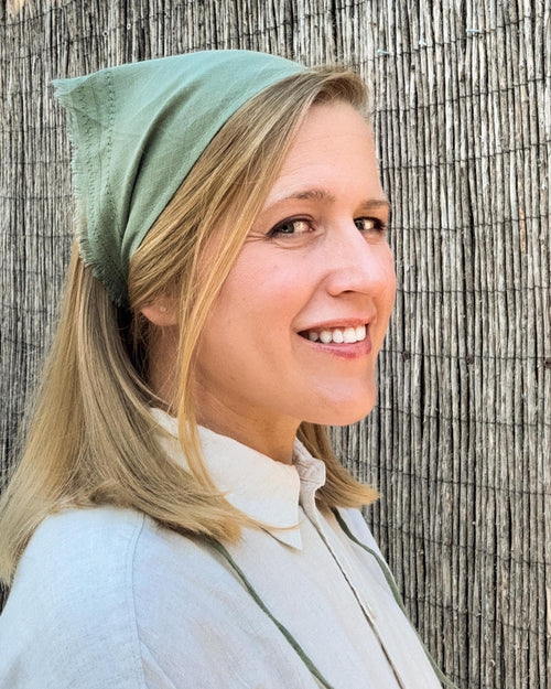 A smiling blonde woman wearing a sage green fabric head covering or bandana, paired with a light-colored collared shirt, standing against a textured bamboo or reed screen.
