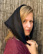 A woman wearing a dark navy blue fringed shawl over her head and a burgundy top, posing in front of a rustic wooden slat wall.