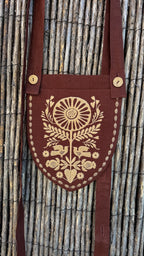 A dark brown linen shoulder bag with a shield-shaped front pocket featuring a beige embroidered sunflower, birds, and floral motifs, shown against a rustic wicker background.