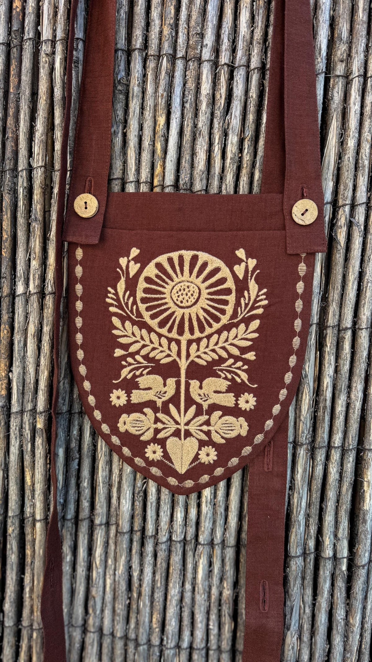 A dark brown linen shoulder bag with a shield-shaped front pocket featuring a beige embroidered sunflower, birds, and floral motifs, shown against a rustic wicker background.