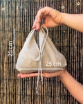 A beige linen drawstring pouch held in two hands against a bamboo screen background, with white dimension lines and text indicating a 25 cm height and 25 cm width.