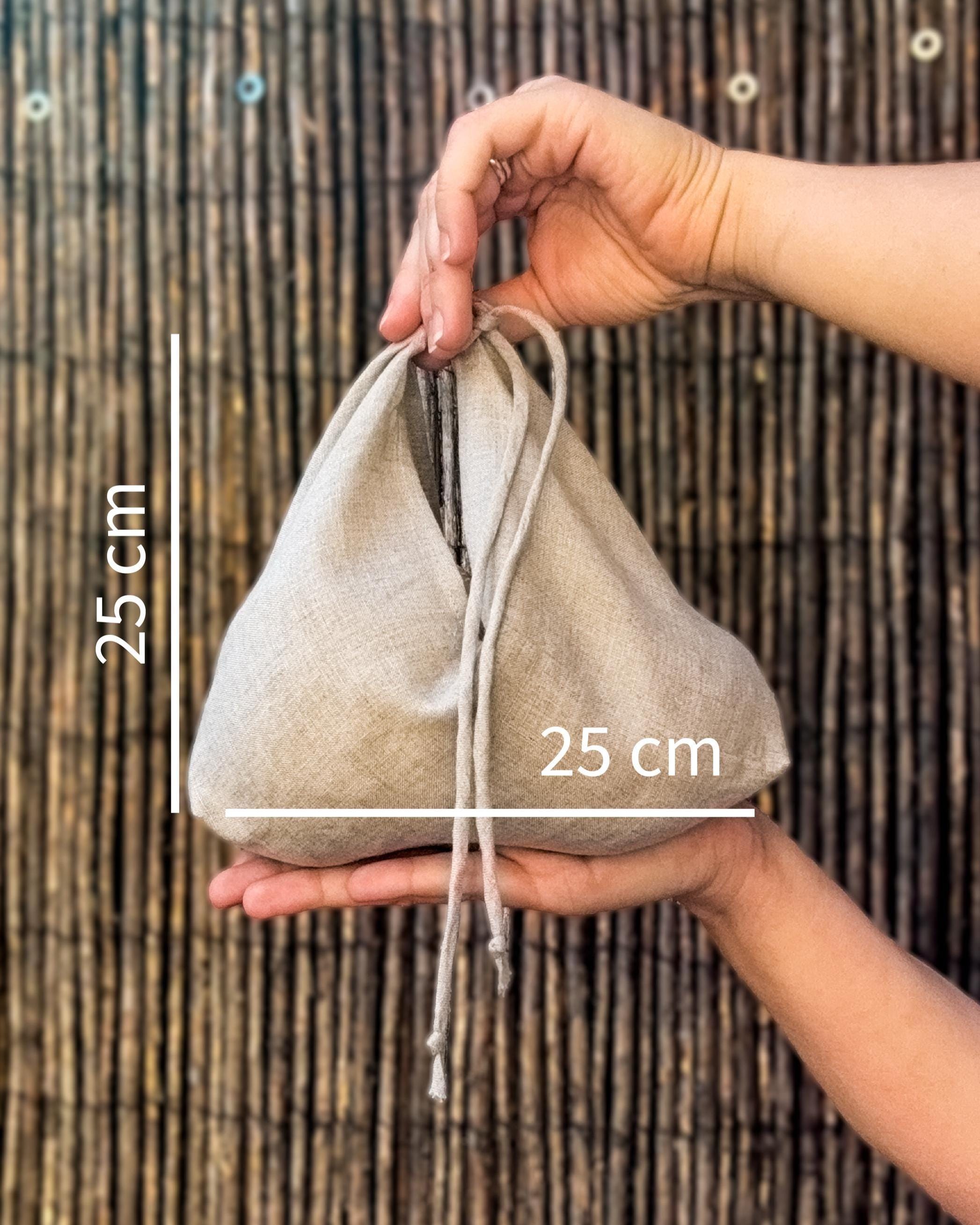A beige linen drawstring pouch held in two hands against a bamboo screen background, with white dimension lines and text indicating a 25 cm height and 25 cm width.