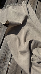 A light beige linen scarf with fringed edges draped over weathered gray wooden planks.