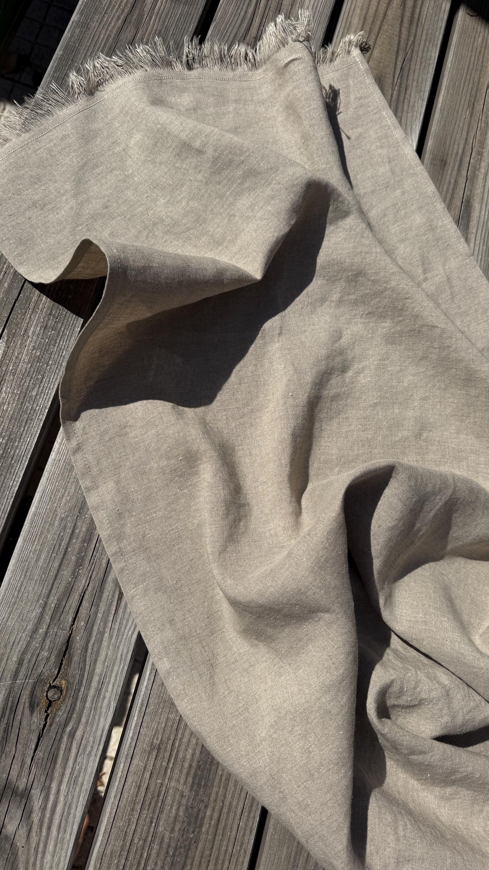 A light beige linen scarf with fringed edges draped over weathered gray wooden planks.