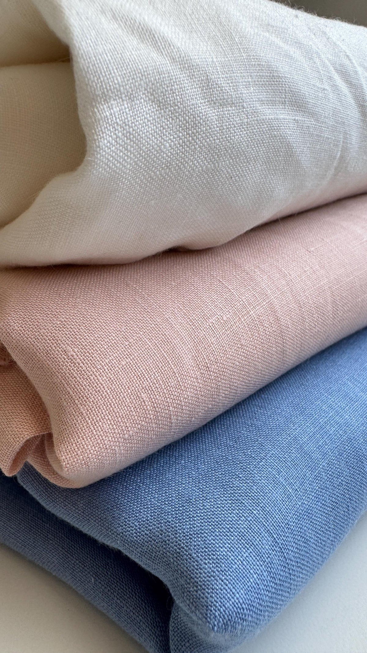Three folded textured fabrics stacked diagonally, showing off-white, dusty pink, and light blue colors.