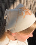 A woman wearing a light beige linen bonnet with orange embroidered floral patterns and fringe trim, photographed outdoors against a bamboo screen.