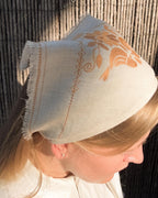 A woman wearing a light beige linen bonnet with orange embroidered floral patterns and fringe trim, photographed outdoors against a bamboo screen.