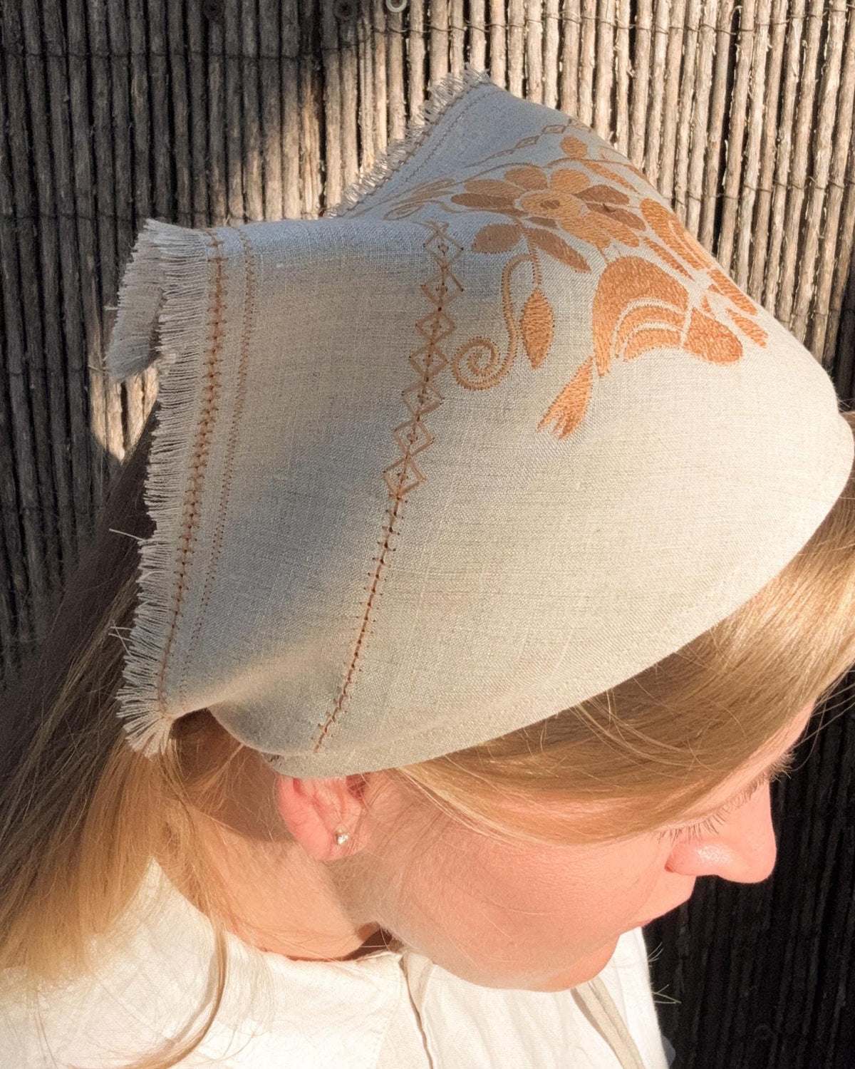A woman wearing a light beige linen bonnet with orange embroidered floral patterns and fringe trim, photographed outdoors against a bamboo screen.