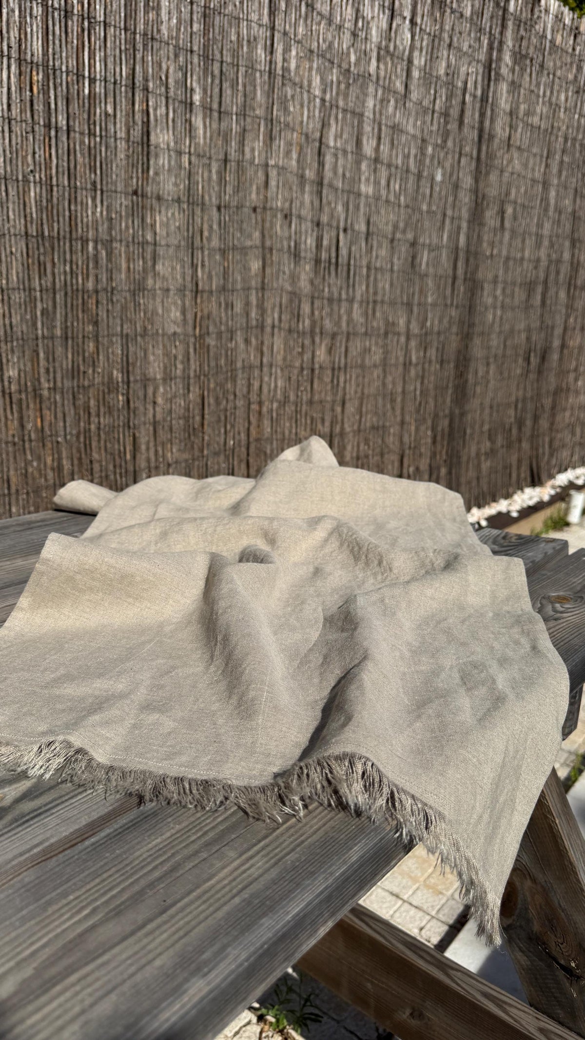 A light beige linen throw blanket with fringed edges draped over a dark wooden table, set against a rustic brown woven reed fence background.