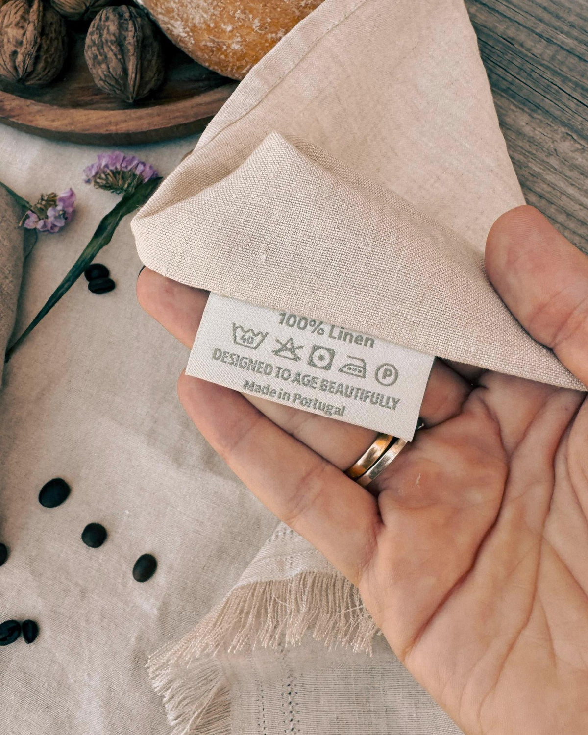 A close-up of a hand holding a folded beige linen cloth, showing the fabric tag that reads '100% Linen' and 'Designed to Age Beautifully Made in Portugal'. The background includes a rustic wooden surface, scattered walnut shells, and dried flowers.