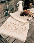 Embroidered linen table runner in natural Belgian linen with wheat design, set on a wooden table with bread and fruit