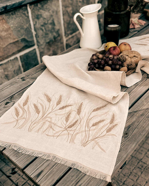 Embroidered linen table runner in natural Belgian linen with wheat design, set on a wooden table with bread and fruit